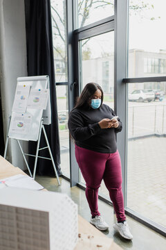 african american plus size businesswoman in medical mask using smartphone near blurred house model.