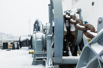 The mooring deck of a cargo ship with anchor winches, an anchor chain on winches, bollards and haws with mooring ropes, frozen and covered in snow. Snow on the mooring deck of a cargo ship.