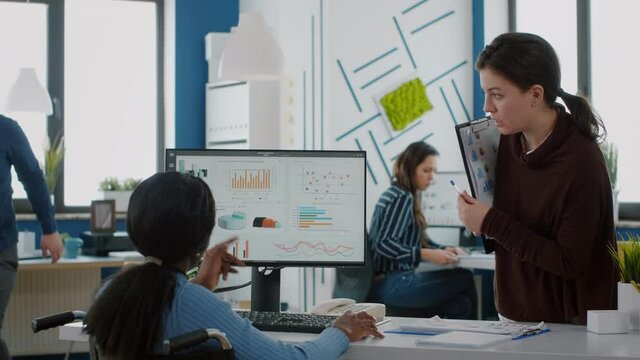 Businesswomen planning financial strategy looking on computer working together in start up company office, black paralysed woman in wheelchair and coworker discussing solution for project.