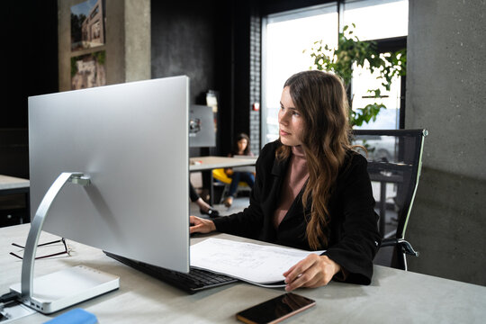 Young woman architect working with blueprints and technical design project at work table and computer in office. Designer using architectural plans of buildings, working at computer, creates project