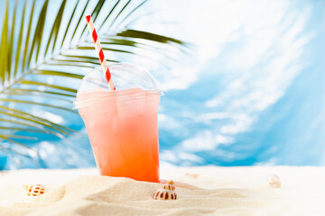 Fresh cold strawberry cocktail in transparent misted plastic glass with straw, ice on sunny beach with palm leaves, ocean and white sand, copy space.