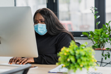african american plus size businesswoman in medical mask typing on keyboard near computer monitor.