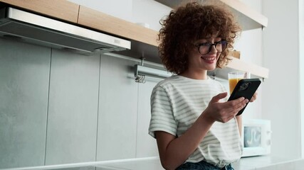 A laughing curly-haired girl in an eyeglasses looking at the phone and to the side with a cup of juice in the kitchen - Powered by Adobe