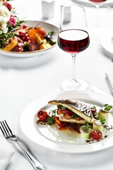 grilled sea bass with sauteed tomatoes, served with a potato ball on a white plate, sea bass fillet plan on a light background, grilled fish fillet served with a glass of wine