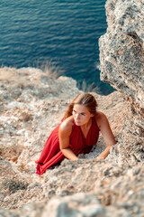 A girl with loose hair in a red dress stands on a rock rock above the sea. In the background, the sea and the rocks. The concept of travel