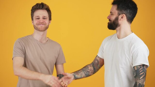 Two handsome guys are shaking hands standing isolated over yellow wall in the studio