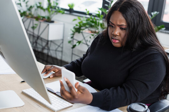 African American Plus Size Businesswoman Looking At Smartphone Near Computer In Office.