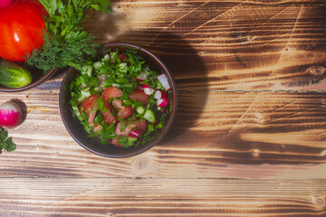 Salad of fresh spring vegetables in a clay bowl. Salad ingredients radish, tomato, cucumber, onion, parsley, dill on a brown wooden table