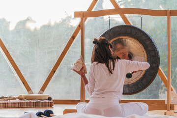 Spiritual woman hitting gong while calm people lying on yoga mats with eyes closed in yoga class