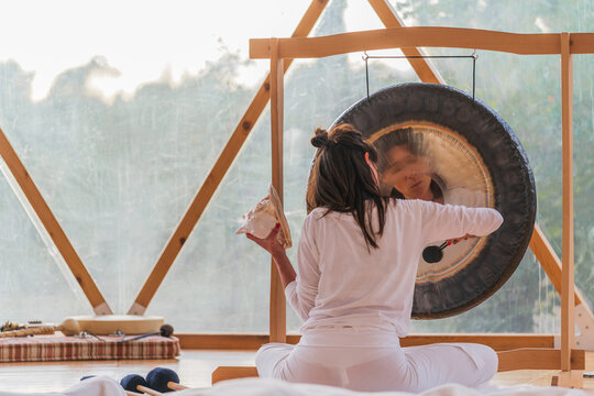 Spiritual woman hitting gong while calm people lying on yoga mats with eyes closed in yoga class