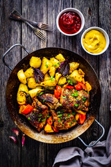 Tasty roasted ribs with baked potatoes and  vegetables served on frying pan on wooden table
