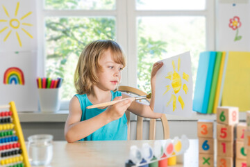 Funny beautiful happy child boy draws laughing with paint. Kid shows drowing of sun. Selective focus.