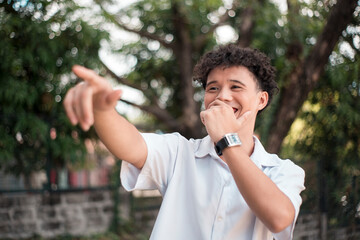 An young mixed race man points to someone, laughing out loud. Comedic situtation or mocking a person. Outdoor background. - obrazy, fototapety, plakaty