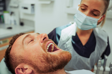 Caucasian male sitting in chair with open mouth while female nurse checks teeth fillings 