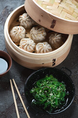 Asian soup dumplings in a bamboo steamer and seaweed salad, vertical shot on a brown stone background, selective focus