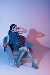 pleased young woman holding sunglasses and magazine while sitting in armchair on purple.