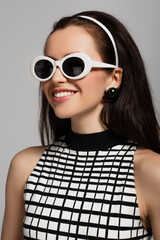 trendy young model in sunglasses and headband smiling isolated on grey.