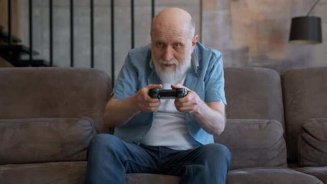 Excited funny gray-haired elderly gamer holding controllers is playing video game on console. Happy pensioner who has won video game reacts emotionally to winning or losing video game. winner in game.