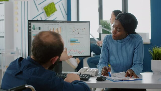 Paralysed invalid entrepreneur in wheelchair and african employee working at financial project for start up company, analysing statistics looking at computer finding solution for business evolution