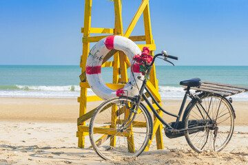 Fototapeta premium Old retro black bicycle with flowers bouquet beside Life ring for life safety on yellow lifeguard stand station or lifeguard chair protecting the safety of tourist on the empty beach in sunshine.