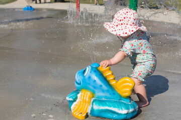 One year old toddler wearing sun protective hat and swim suit at a splash pad water park; baby...