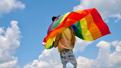 Blonde woman holding a rainbow LGBT gender identity flag on sky background with clouds without face in a month of dignity of LGBT Communite