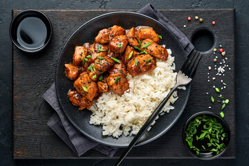 Spicy teriyaki chicken fillet pieces with rice, green onions and black sesame seeds on black plate on a dark slate, stone or concrete background. Top view with copy space.