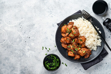 Spicy teriyaki chicken fillet pieces with rice, green onions and black sesame seeds on black plate on a grey slate, stone or concrete background. Top view with copy space.