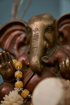 A close up of a statue depicting Oshiro Ganesh with prayer beads and a shamanic drum with teal background. warm tones contrast with cool colors with a lot of detail and interest.