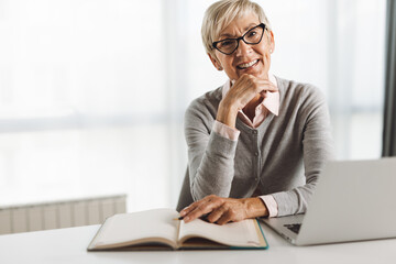 Happy senior businesswoman sitting in the office and using laptop while looking at camera