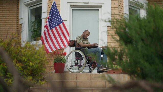 Disabled African American military man in wheelchair looking through pictures smoking cigarette on backyard porch. Wide shot of paralyzed paraplegic sad frustrated veteran soldier recalling memories