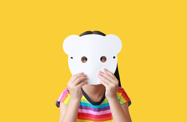 Little child girl holding blank white animal paper mask fronting her face isolated on yellow...