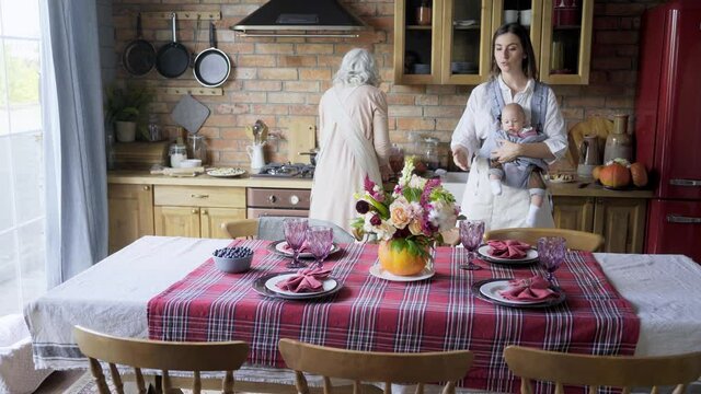 ladies serving and prepare large table to celebration, Young woman carry baby in sling bag and helps aged mother set glasses, plates with food and festive pie on checkered tablecloth 
