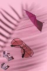 pink sunglasses, beach umbrella and ice cubes under a palm tree on a pastel background. hard shadows from the sun at noon. leisure, travel and entertainment concept with copy space