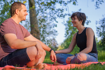 Photo of a young and attractive couple sitting on a towel in the park  smiling at each other and talking. Great summer day, relaxed attitude.