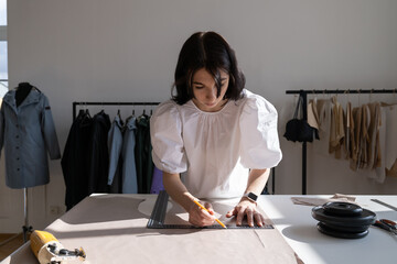 Female seamstress draw patterns for cutting, measure material on big table in atelier. Garment...