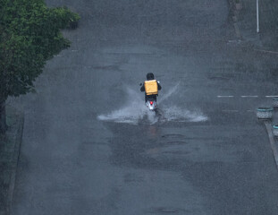 Food delivery courier with a yellow box on his back delivers an order to a customer on a scooter on a rain-flooded road. Driving through puddles.