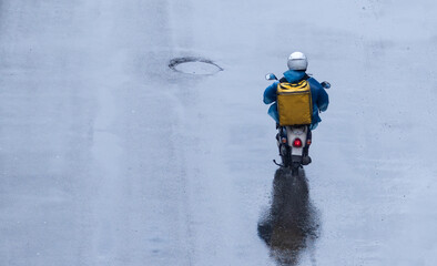 food delivery man in a blue raincoat with a yellow box on his back is eating on wet asphalt