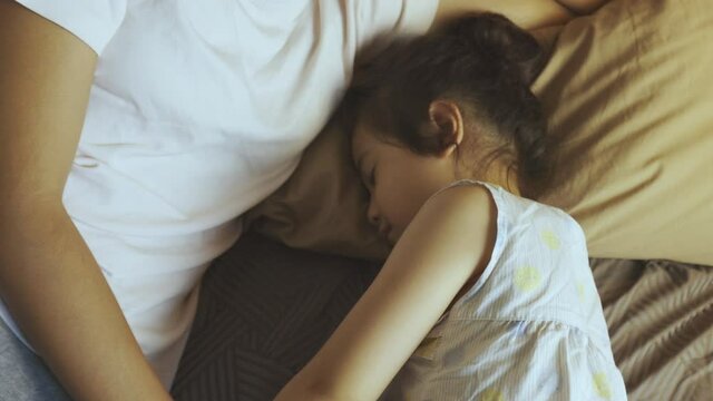 Adorable 5 years' old asian girl is sleeping on bed in mother's hold to listening bedtime story and spending good family relation moment which shows relax, enjoy, happy and positive togetherness.