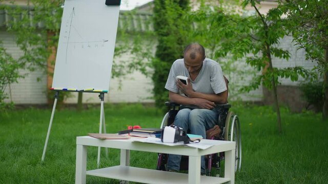 Depressed paralyzed African American man hugging photo sitting in wheelchair in summer garden. Portrait of desperate frustrated guy regrets injury outdoors on remote working