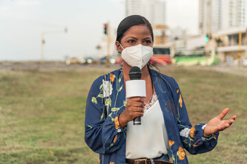 African American woman reporter in mask speaking into microphone while standing on street in city