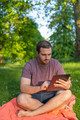 Photo of a young and attractive man sitting in the park on a towel enjoying a sunny day outside in the nature and checking his tablet. Electronic device, technology
