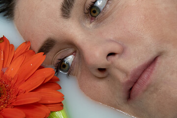 Woman in a milk bath with her eye partway in the water looking at orange flower petals.. Her eyes, lips, nose, and eyebrows can be seen.