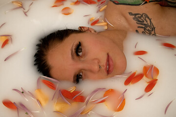 Tattooed woman in a white milk bath with orange flower petals. She has eyeliner and mascara on which surround her brown eyes. Only her head and shoulder can be seen.