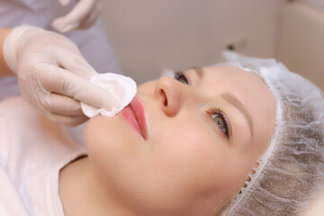 The cosmetologist wipes the remnants of anesthesia from the client's lips with a cotton sponge