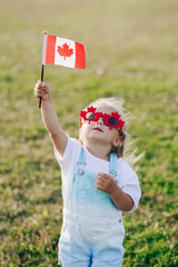 Happy Canada Day. Little Caucasian toddler girl in funny maple leaf sunglasses waving Canadian...