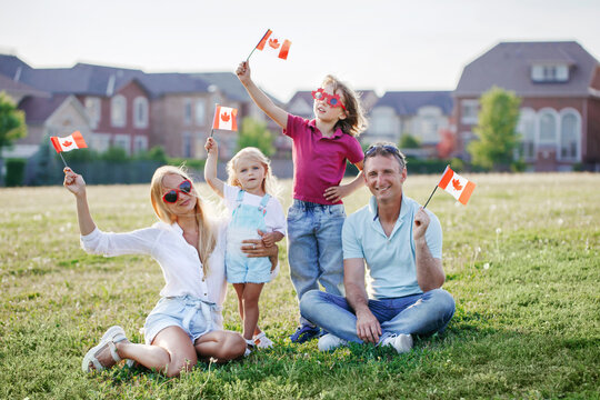 Happy Canada Day. Caucasian family with kids boy and girl sitting on ground grass in park and waving Canadian flags. Parents with kids children celebrating Canada Day on 1st of July outdoor