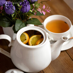 Tea ceremony. Full ceramic teapot of brewed black tea with orange slices and tea cup. White ceramic utensils on wooden table and flowers. Close up shot. Soft focus.