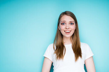 Obraz premium A cheerful girl in a white T-shirt is surprised and happy while standing on a blue background and opening her mouth