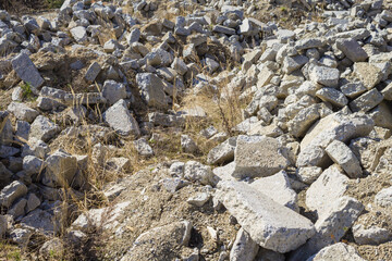 Heaps of dumped, gray, debris of concrete slabs and stones among the yellow, dry grass. Landfill for construction waste. Traces of an earthquake and a devastating catastrophe. Desolate area.